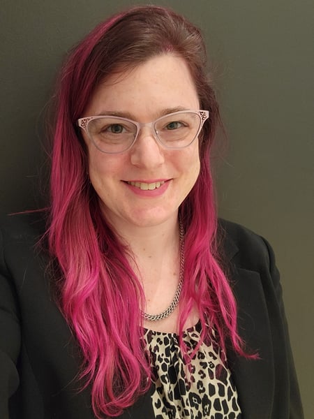 Meredith smiles in a portrait, wearing a blazer and shell top. Her hair is bright pink and her glasses are soft pink.