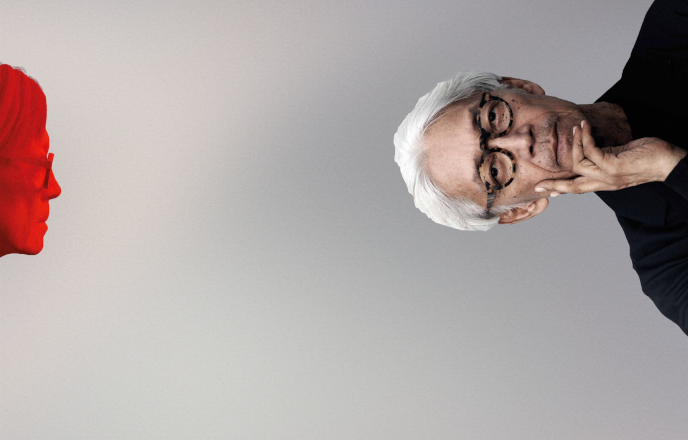 Two images of Japanese composer Ryuichi Sakamoto on a grey background; one of him in profile on the left hand side, his face coloured red; and one on the right hand side, oriented sideways, with white hair, round glasses and his left hand cradling his cheek and chin.