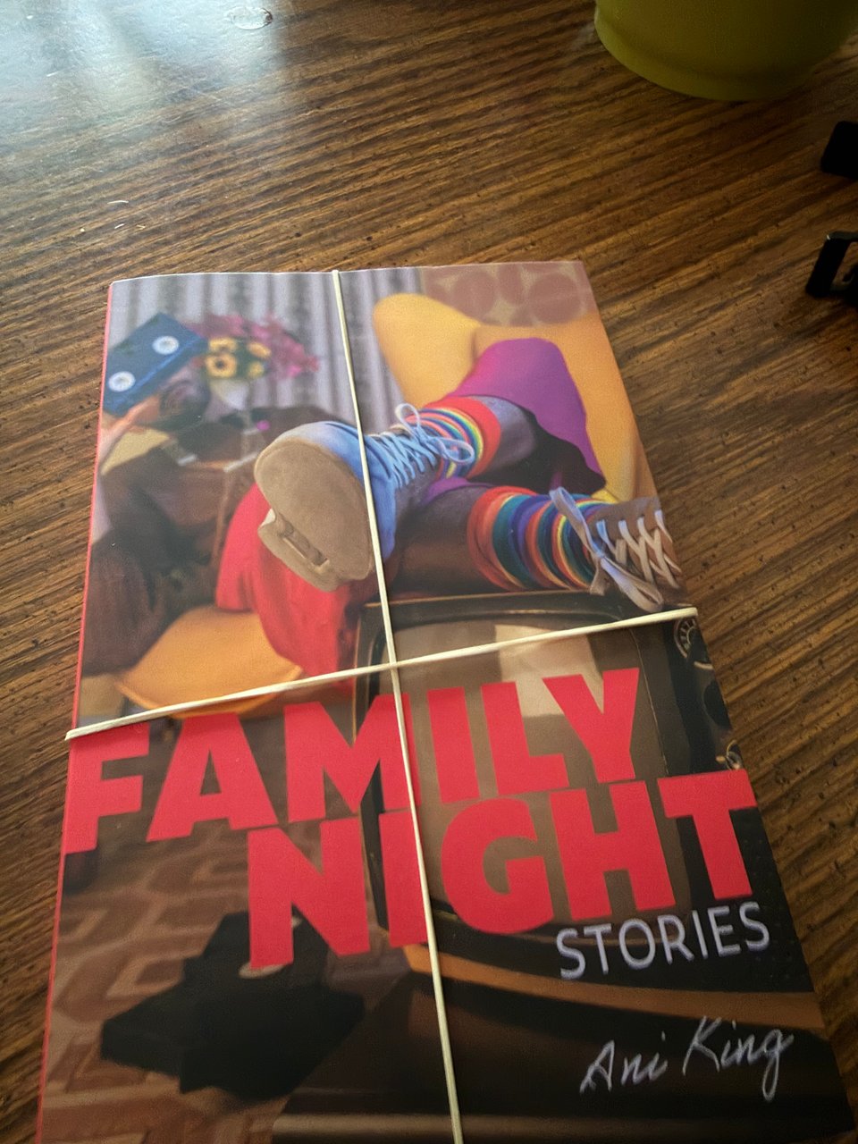 Picture of Family Night: Stories by Ani King bound in two tan rubber bands and sitting on a wooden table. The Family Night official book cover features a person with a mustache and short beard reclining in a yellow chair and holding a VHS tape over the top of their face so the white spool pieces look like eyes. They are wearing rainbow striped socks and lace up shoes, with their feet resting on a vintage television set. A small pile of VHS tapes rests on the floor, and FAMILY NIGHT stretches across the cover image in bright pink capitals, with Stories and Ani King in underneath in smaller, light blue font.
