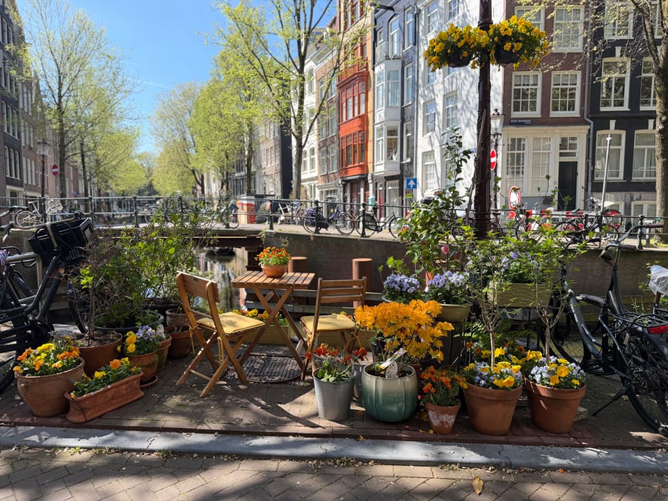 A canal side street in Amsterdam. The sidewalk is filled with flowers and plants and a table and two chairs.