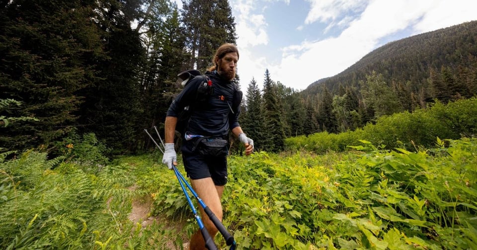 A man with a backpack and trekking poles walks along a narrow dirt path surrounded by lush green vegetation. He is in a forested mountain area under partly cloudy skies.