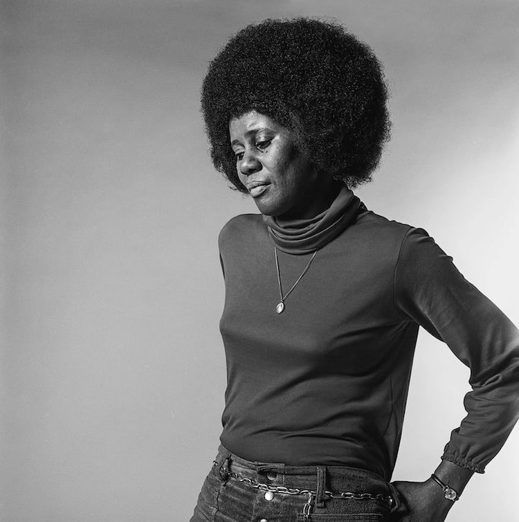 A Black woman looking down. She has an afro and is wearing a rollneck top and jeans with a chain belt.