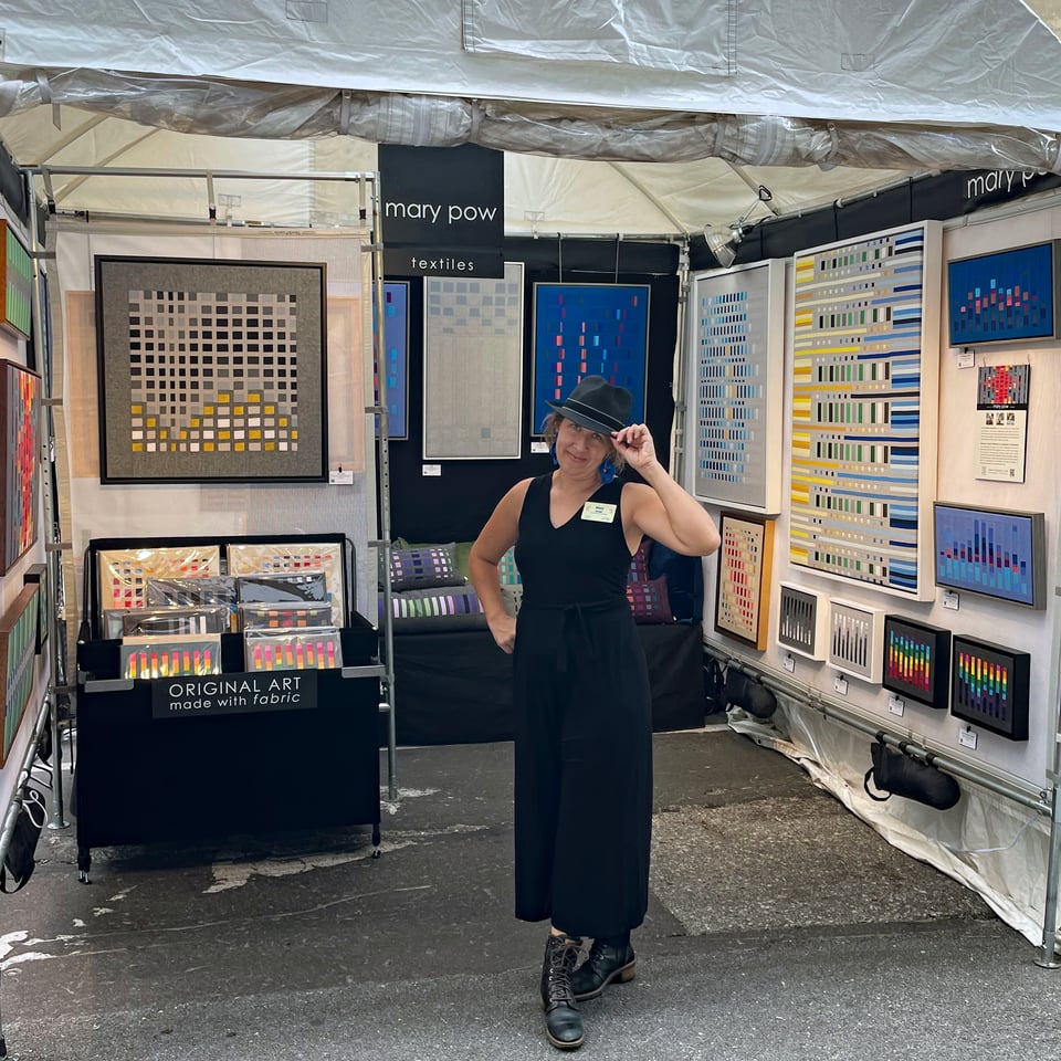 A photo of Mary Pow tipping her hat in front of her textile art booth at an art fair.
