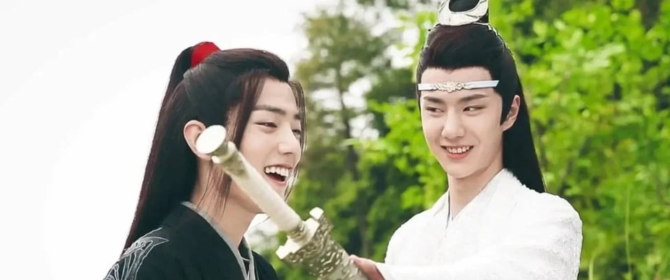 Photo of Xiao Zhan and Wang Yibo in costume on the set of The Untamed in 2018. They are both smiling as Yibo holds his character's sword playfully in front of Xiao Zhan.