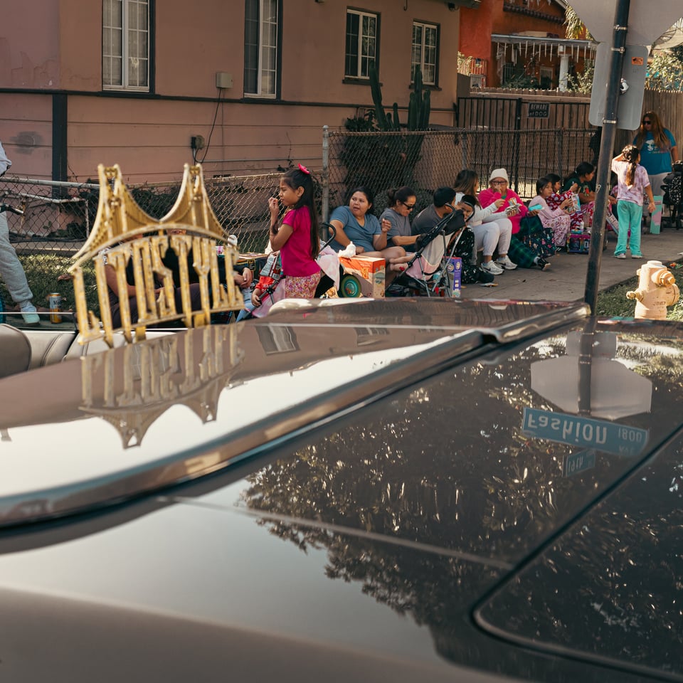Photo of various people in a Long Beach community seated on the sidewalk eating food, while in the foreground is a metal plaque with the word "LITOSTYLE," in reference to the lowrider car club by that name.