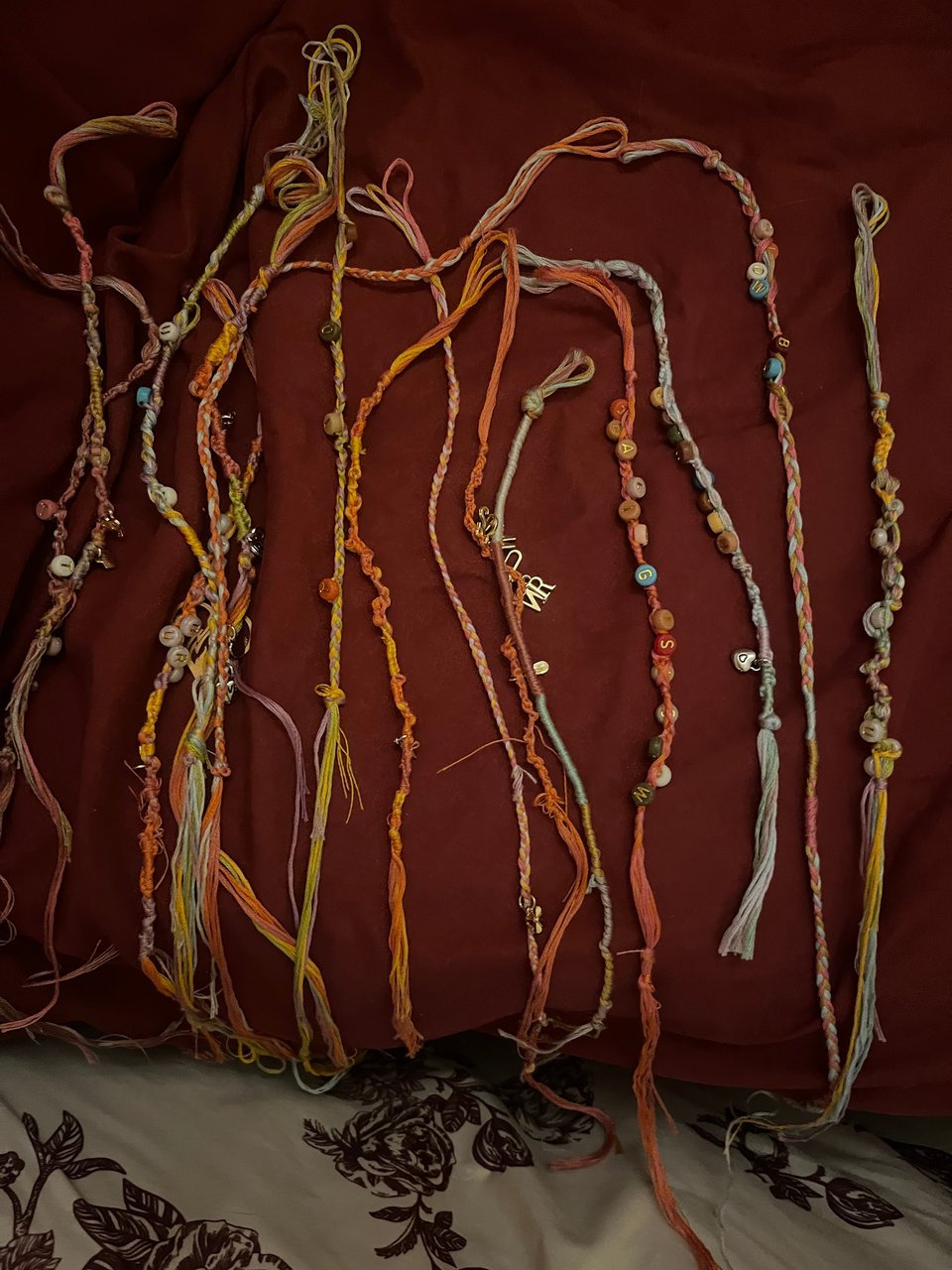Several handmade friendship bracelets against a dusty pink couch with white fabric with maroon rose print in the foreground. Bracelets are in a range of colors - gold, orange, light blue, lavender, and pink with a range of beads and charms.
