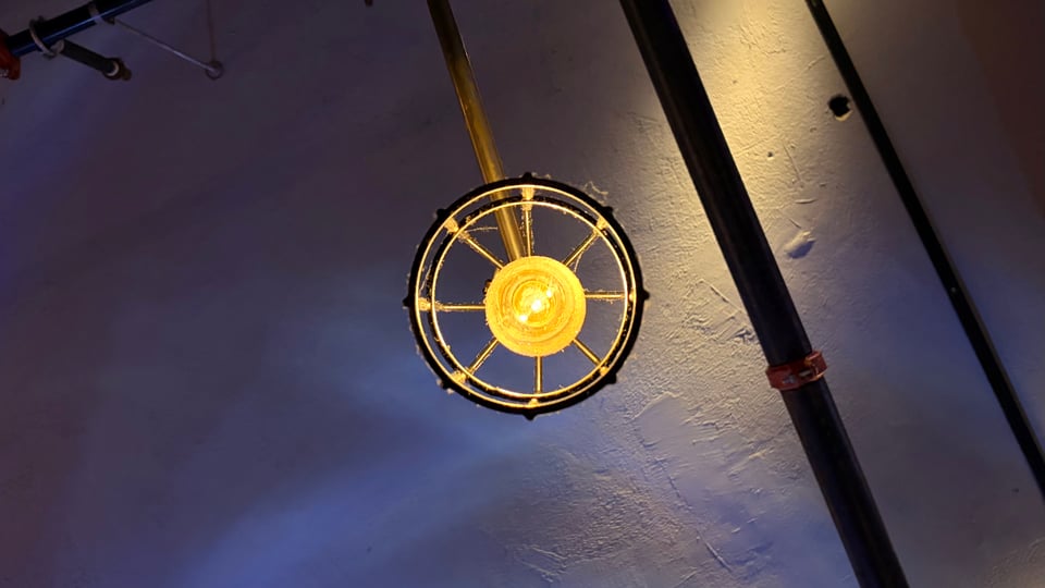 View from below of a very yellow lightbulb in a lamp that looks like a metal cage, which is dusty and partly covered in cobwebs, held up by a metal tube. In the background, an industrial-looking ceiling with more metal tubes, lightly tinted purple by other ambient lighting