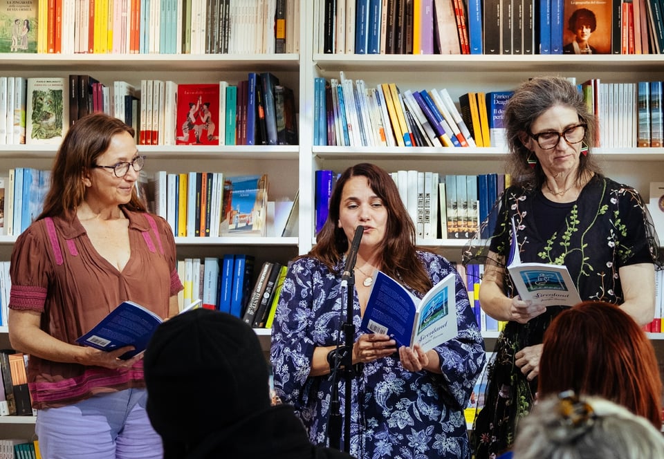 Alt text: Three people holding the same book stand in front of a bookcase, as one reads in to a microphone