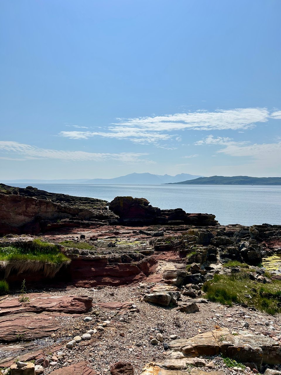 A beach full of craggy reddish rocks, with hazy island mountain peaks in the distance. The sea is calm and pale blue and the sky above is brighter blue with wispy and puffy white clouds. Image by Rowan Ambrose..