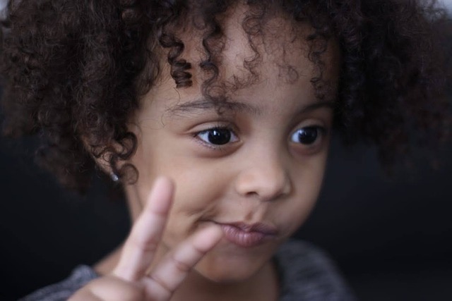 toddler-aged black girl looking down and to the right of the camera, holding a peace sign, but her mouth is twisted as in "I guess so"