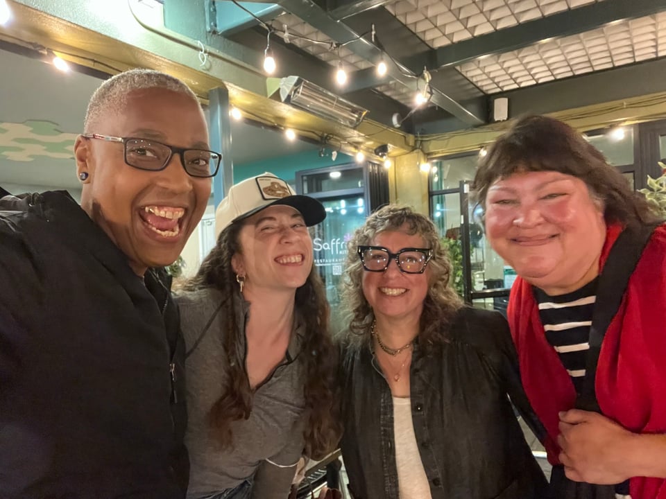 Saifa and Hans smiling in a goofy group picture at an outdoor restaurant. Next to Hans is Thea, a white intersex woman with curly gray hair and thick rimmed black glasses, and Niki, a mixed race intersex woman in a red cardigan.