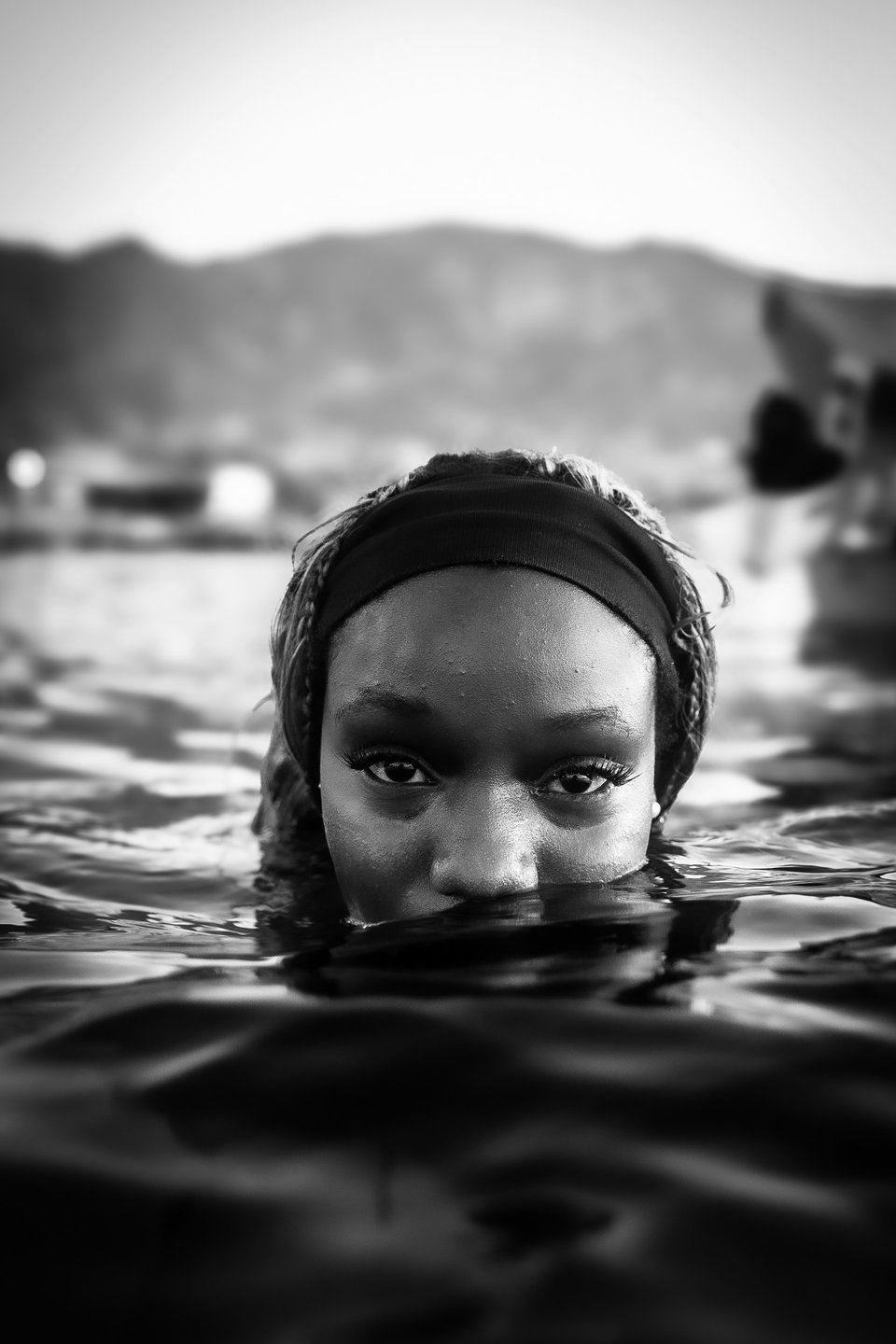 Black and white photo of a woman rising from the ocean. The water covers her mouth and most of her nose and she's staring directly at the camera. Out of focus in the background is some buildings and a hill past the beach.