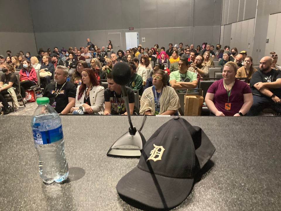 a detroit tigers baseball cap and a bottle of water next to a microphone, looking out at an audience at a comic book convention