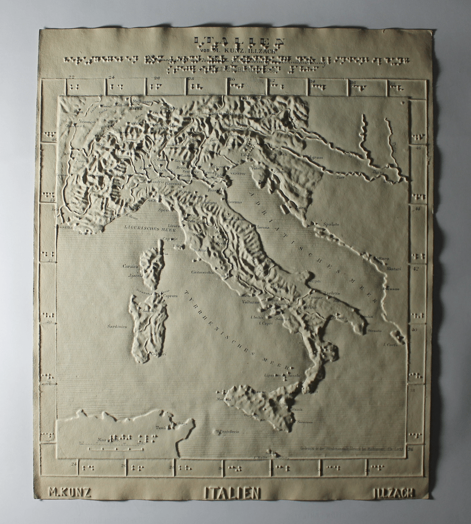 Large piece of thick cardboard with an embossed image of Italy, rendered as a relief map. The top of the map has a few lines of braille. The map looks very old.