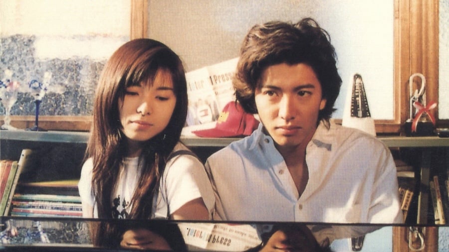 A young man and woman sit closely together at a piano, appearing contemplative in a warmly lit room filled with books and decor.