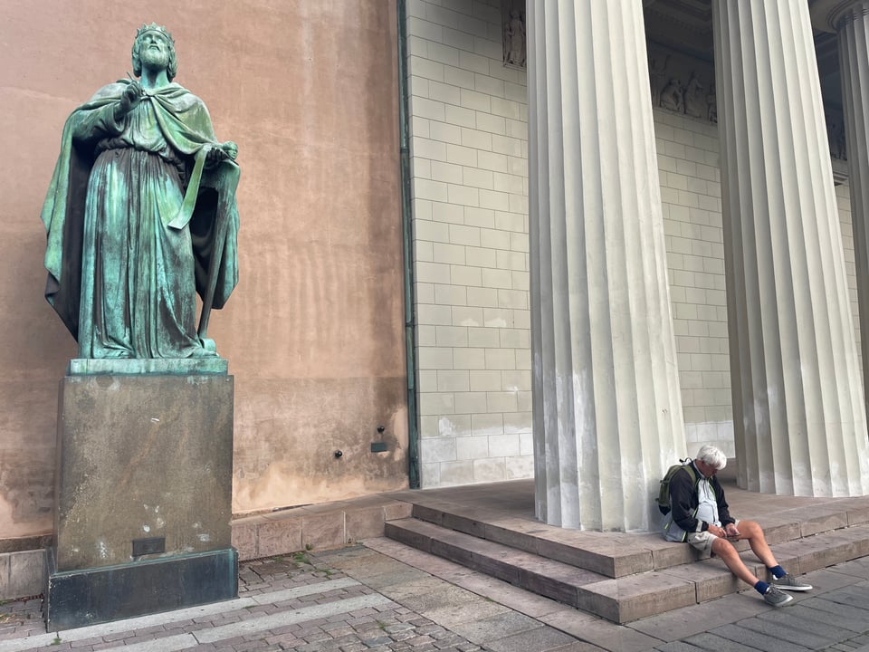 A large green statue shows a crowned man looking up to the sky. Next to him a bit away there is a man sitting on the steps looking down (probably at his phone).