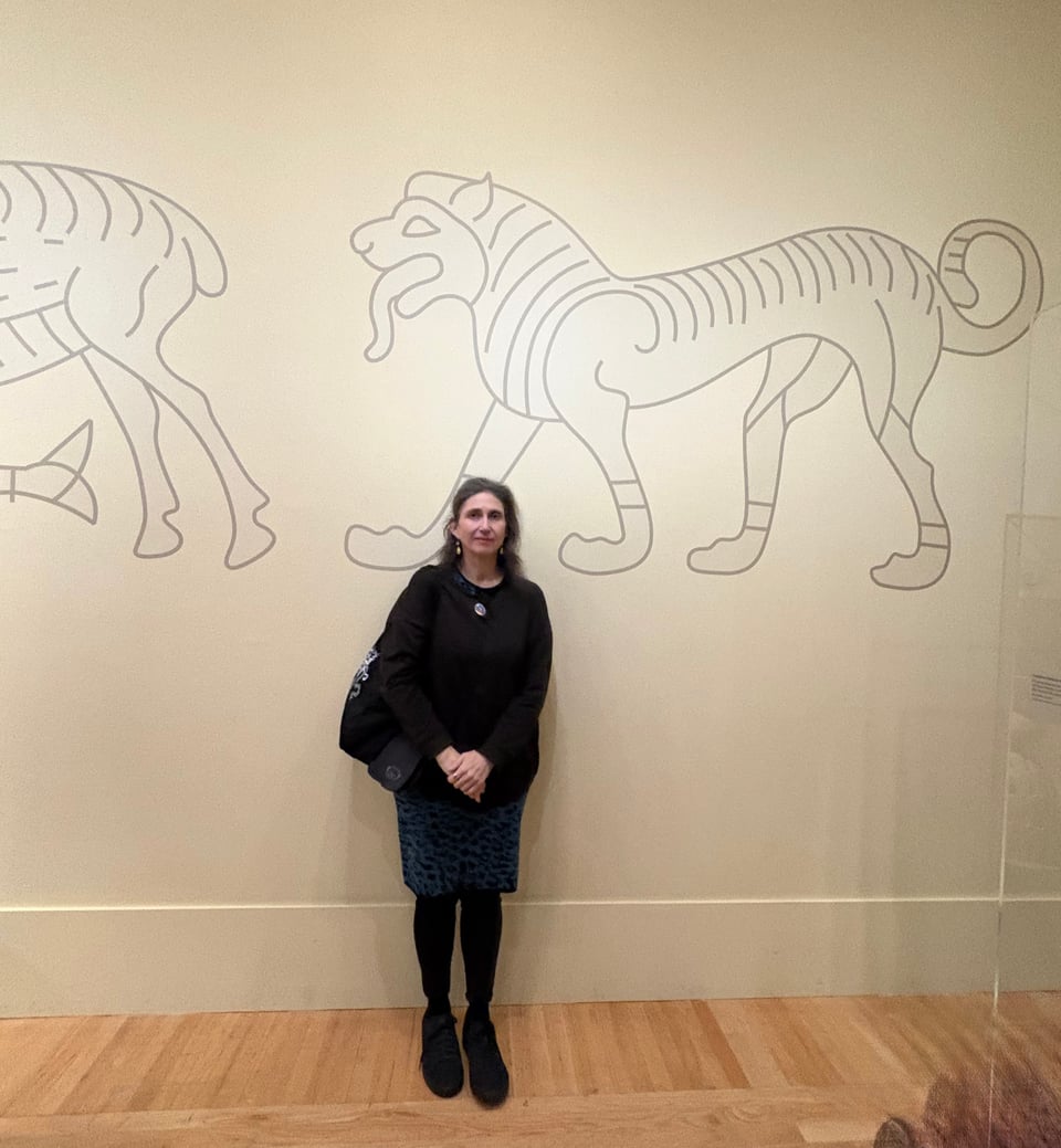 Alt text: A person poses in front of a wall with Etruscan inspired art on it.