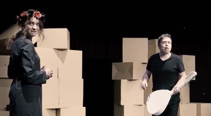 Two performers are on stage in front of stacked cardboard boxes, one wearing a flower crown and a black outfit, and the other holding a stringed instrument.