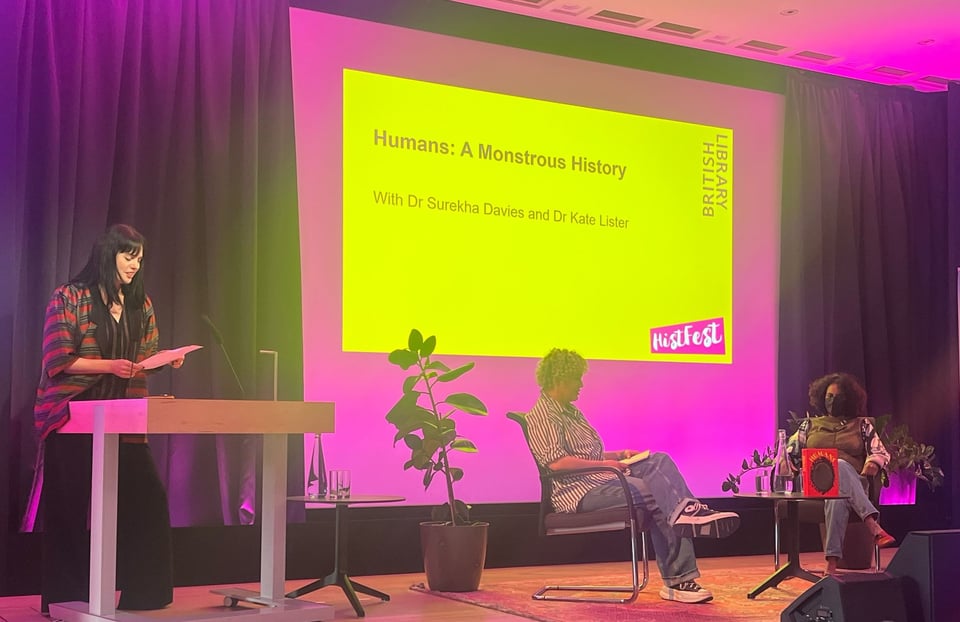 Stage with giant, bright yellow screen with pink border, with the pink and white “HistFest” logo at the bottom right. Two women seated in front of screen, around a small table with a bottle of water, two glasses, and a bright red book with dark oval circle on the cover sits on the table.