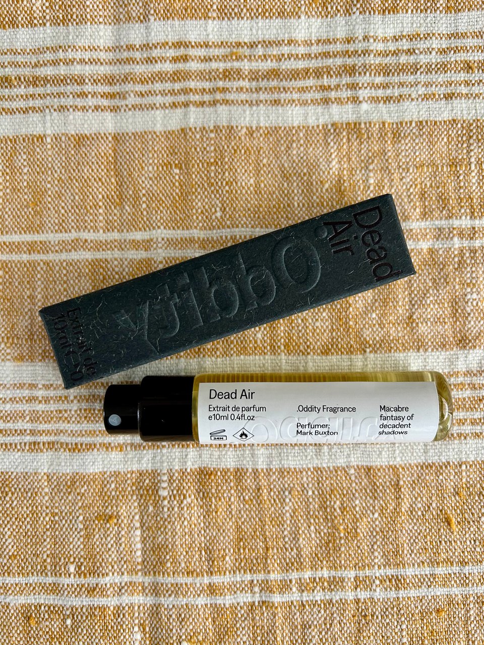 A travel size glass bottle of perfume with Dead Air by Oddity branding is lying next to a dark grey Oddity box on a gold and white linen cloth. Image by Rowan Ambrose