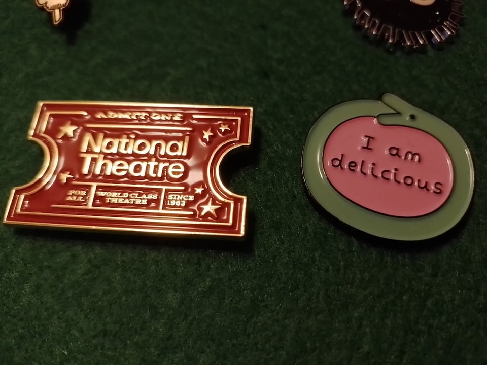 Two shiny enamel pins pinned to a green felt background. One looks like a National Theatre ticket, the other is a cartoon ourobouros saying "I am delicious"