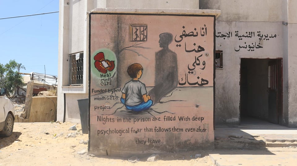A photo showing a painted mural on the wall of a building in Gaza, illustrating a child sitting on a prison cell floor and looking at their tall shadow cast on the wall, like an adult's shadow. There is a barred window with barbed wire above the child, and the text "أنا نصفى هنا وكلي هناك". Below the mural is the text: "Nights in the prison are filled with deep psychological fear that follows them even after they leave." The Heal Minds Gaza logo is at the side.