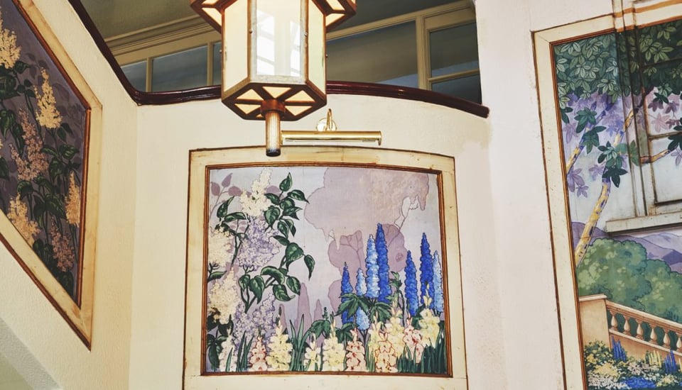 A corner of a foyer area with three murals set into frames on the cream walls. A large hexagonal brass pendant light hangs between them. The murals are in pastels of lilac, cream and blue and feature flowers and trees.