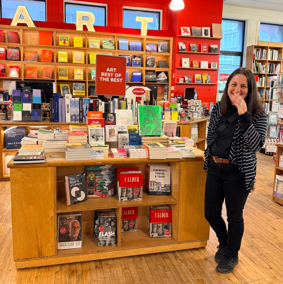 A woman stands in a bookstore in front of a display of books with a bright green book featured in the middle of the table. the table is labeled "art best of the best"