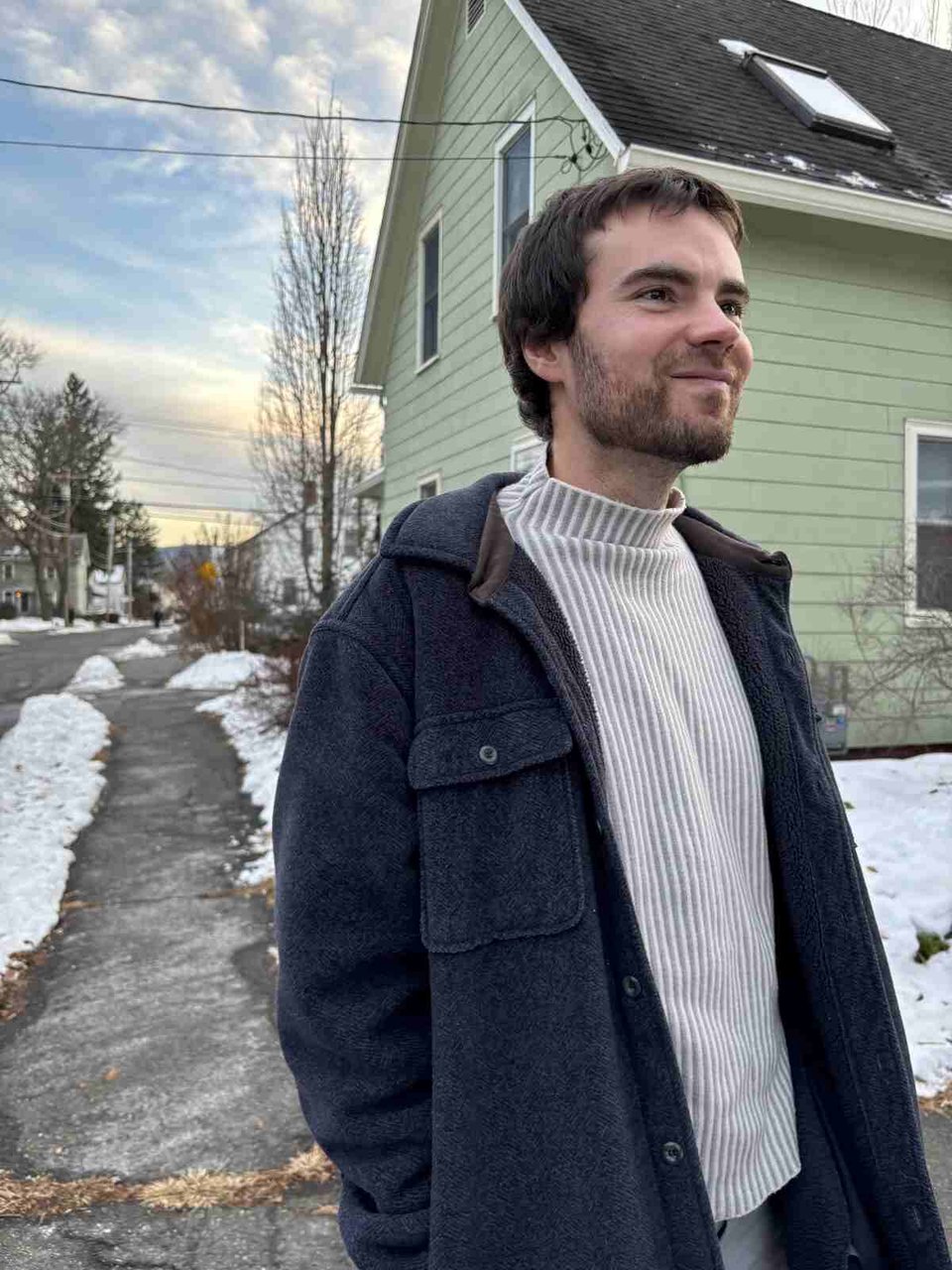 A young man with short brown hair and a short beard wears a dark winter coat over a ribbed white turtleneck. He stands on the sidewalk in front of a green clapboard house, and there's snow behind him.
