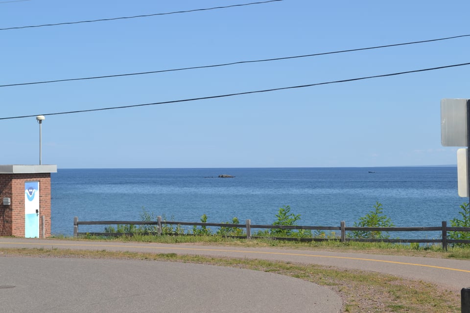A road running along the shore of a large, quiet lake under blue skies.