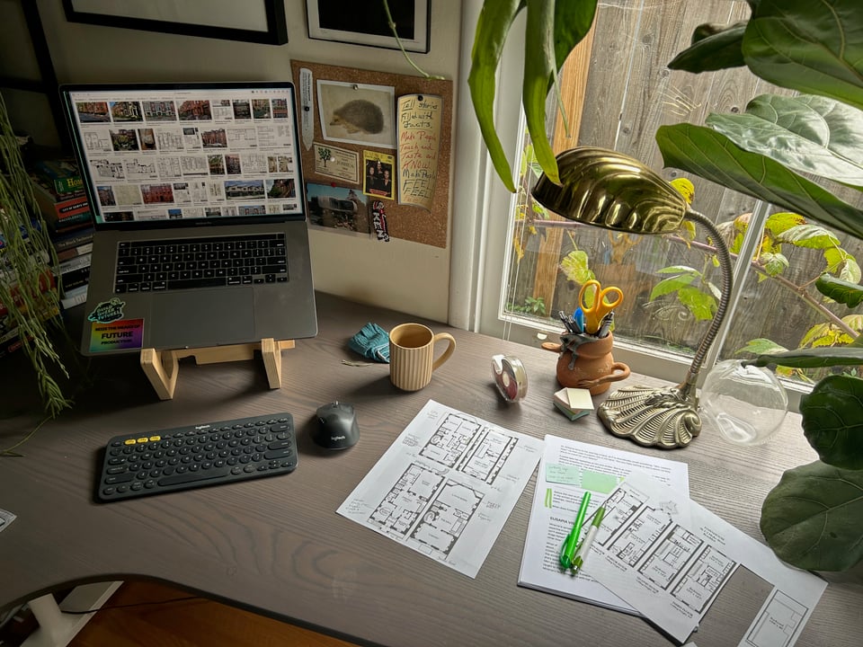 a desk that has both a computer on a stand and a keyboard, as well as a bunch of papers on it, a gold lamp, and several plants creeping in at the edges