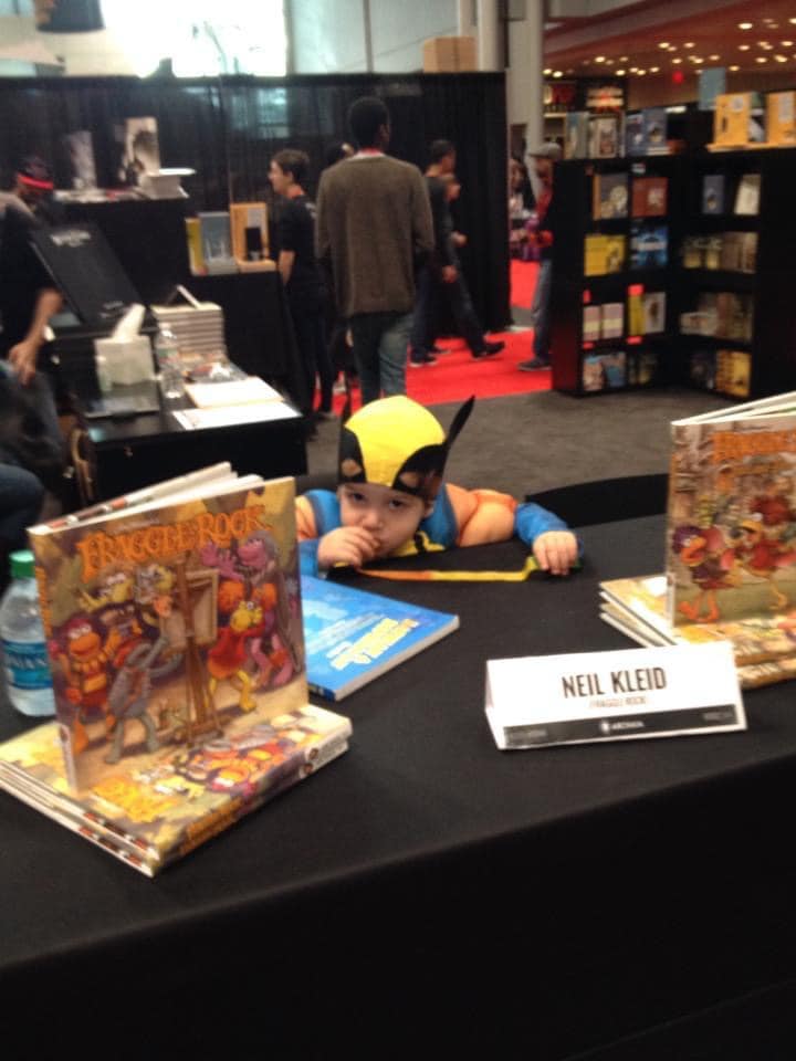 a little wolverine drawing at a comic convention table, with a "neil kleid" placard on a black tablecloth