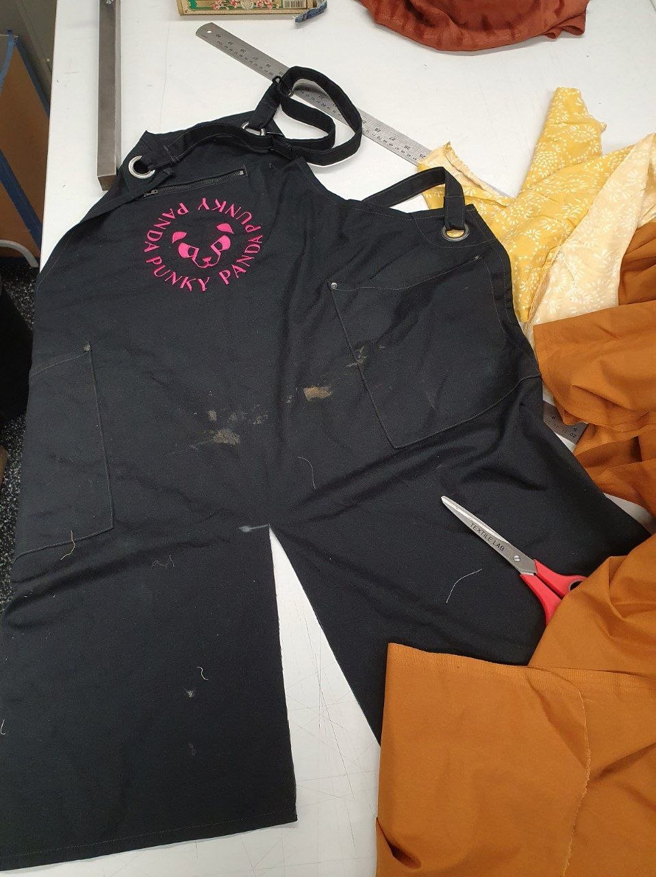 A black apron with a pink logo of a panda on the chest. A cut has already been made, bisecting the bottom half of the apron.