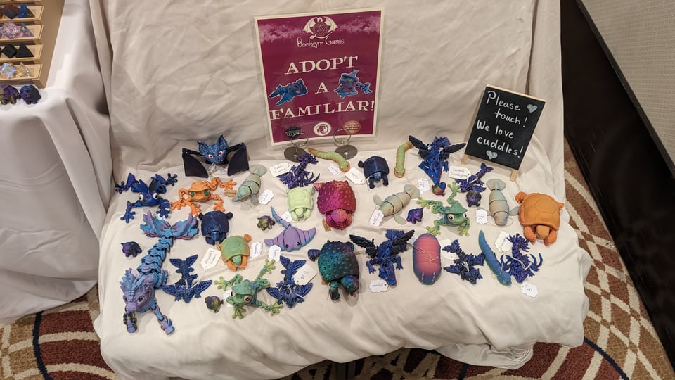 A bunch of cute little 3D printed "familiars" beneath a sign reading "adopt a familiar." A seconed sign reads "Please touch! We love cuddles! <3"