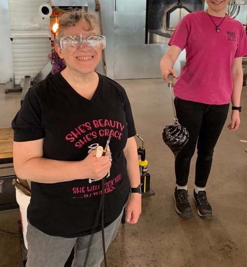 A woman stands at the forefront, grinning, her glasses reflective. She holds tubing for blowing glass in one hand. She is dressed casually in a T-shirt and jeans, and her hair is frizzed and in a ponytail. Behind her a figure in a pink T-shirt and black jeans is cropped to not show a face. The figure holds out a rod with an ornament dangling at the end.