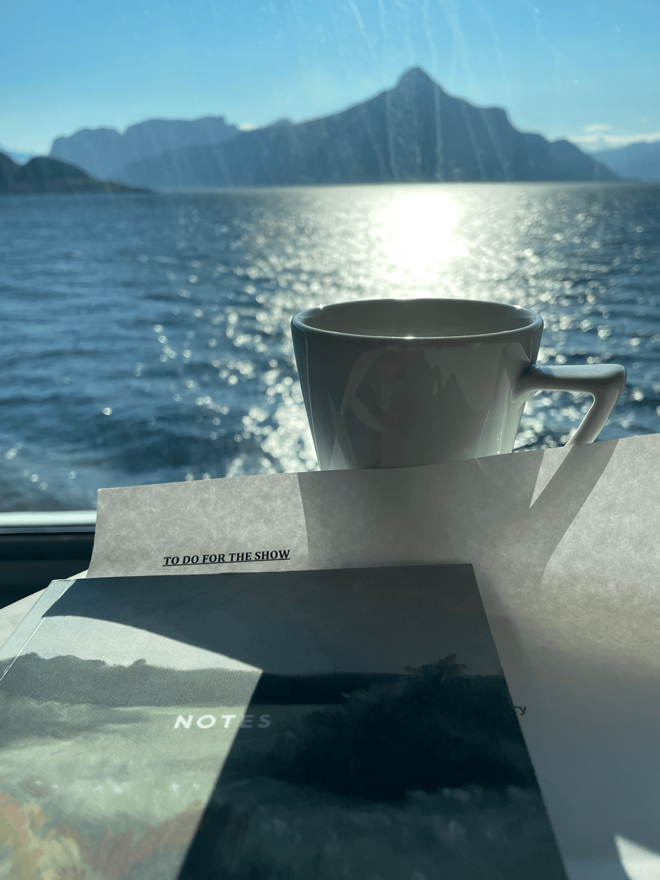 A coffee cup and notes that read "To Do For The Show" with water and mountains in the background