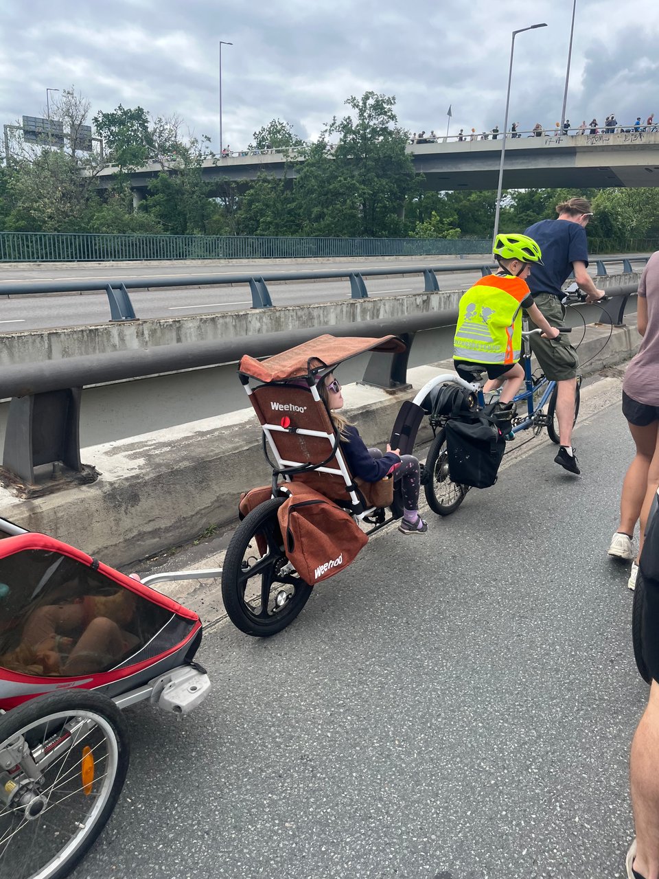 A man on a tandem bicycle with a young child on the second seat. Attached to the tandem bicycle is a cargo recumbrent bike seat with a young girl. Attached to that is a cargo toddler bike trailer.