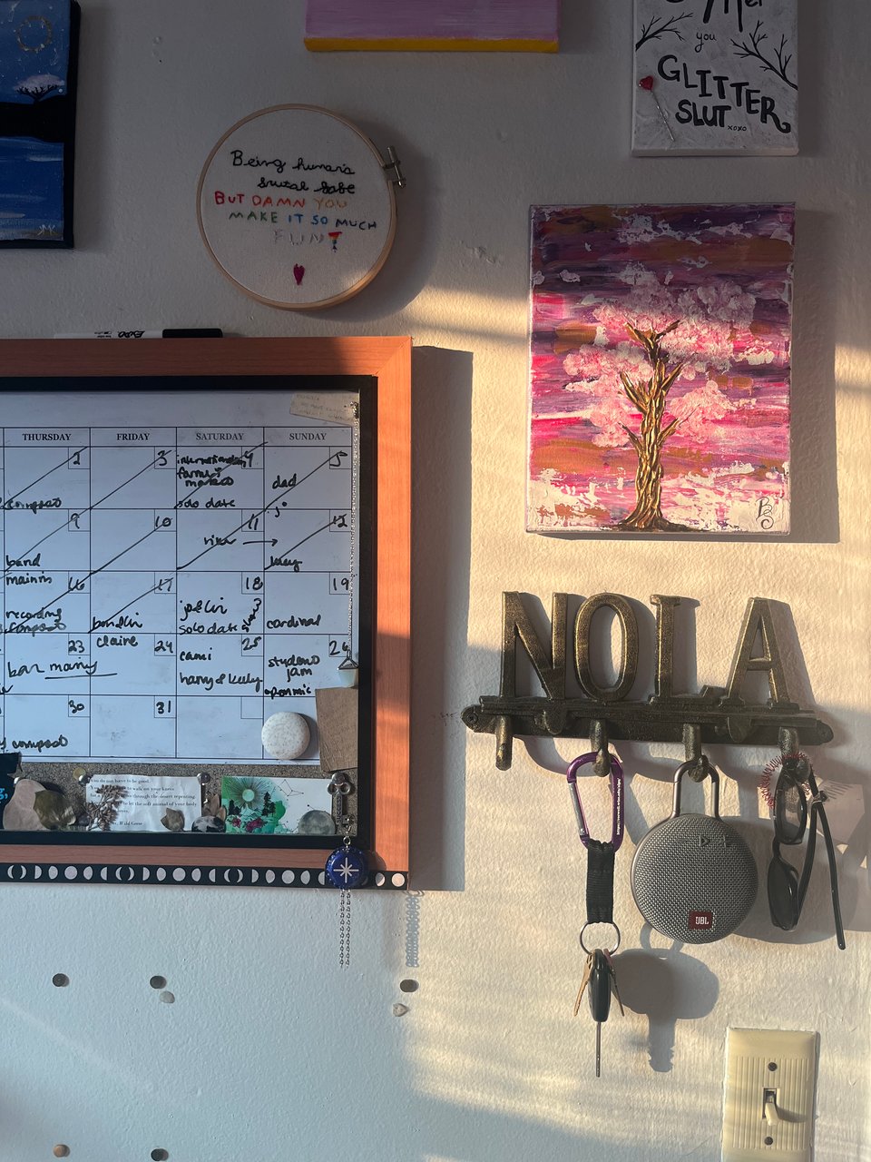 a snapshot of a bedroom wall at golden hour with a whiteboard calendar, friends' paintings and embroidery, and decorative brass hooks that say "Nola"