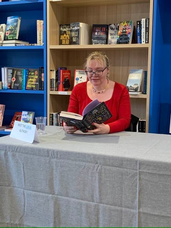 Kate is sitting at a table in a red dress reading from The Valkyrie.