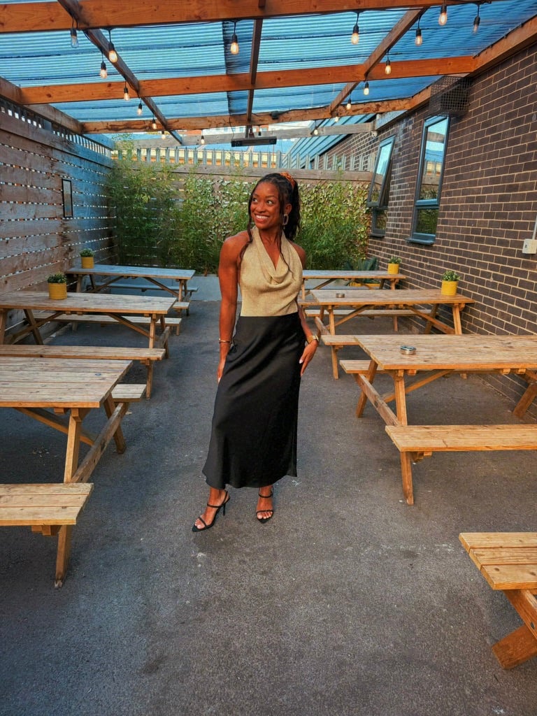 Nadia poses in a covered outdoor seating area, smiling in a gold halter-style top, long black skirt, and black heels.