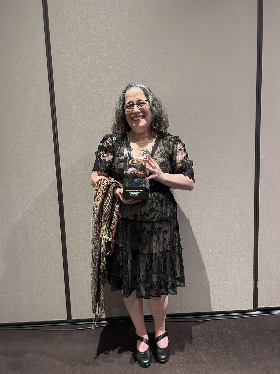 An extremely smiley middle-aged woman with salt-and-pepper shoulder-length hair clutches a Nebula Award in her hot little hands. She's wearing glasses, a black lacy dress, and black shoes, and a plum-and-gold fringed shawl is draped over one arm.