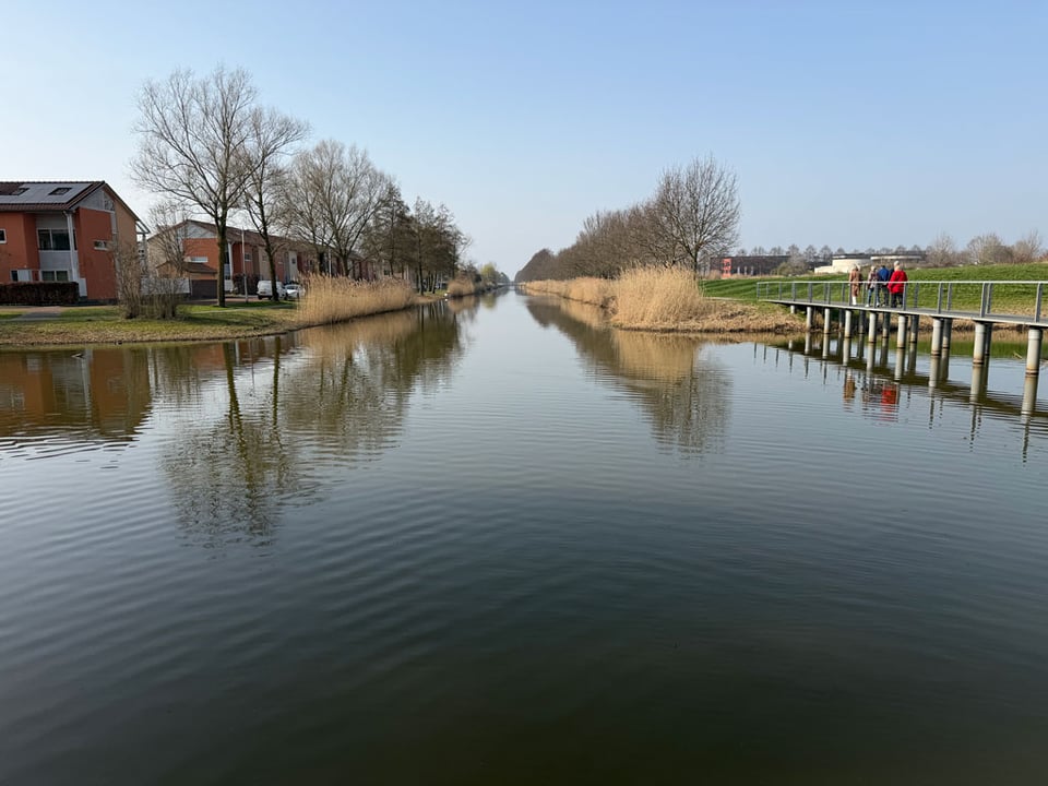 A canal fills up the photo as reed grasses grow on either sides. People walk over a bridge away from the camera.