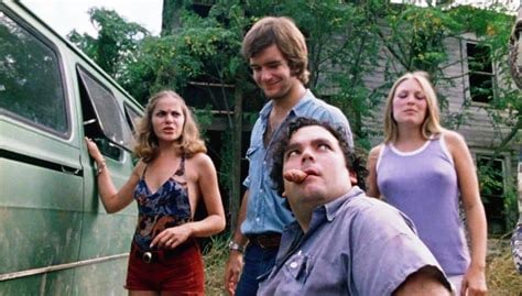 Pam ( a blonde with shoulder length hair ending in a slight curl wearing a dark blue floral halter with red short shorts), Kirk (a brunette with 70s above-the-chin length helmet hair wearing a light blue work shirt opened to the third button and blue jeans held up with a brown leather belt), Franklin (a brunette with short cropped curls in a wheelchair wearing a blue grey work shirt with the top button undone and a piece of sausage held in his mouth), and Sally (a blonde with straight shoulder length hair wearing a purple tank top with white edging and white shorts) stand next to their green van outside Sally and Franklin's rundown former family home.