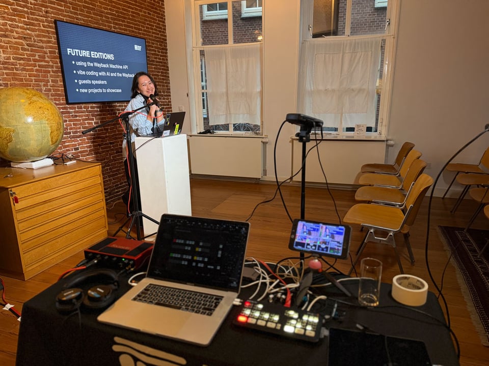 A woman smiles cutely and stands at a podium in a room with empty chairs awaiting to be filled by an audience who has yet to arrive.