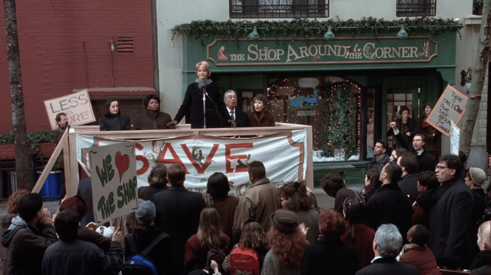 Kathleen Kelly speaking in front Shop Around the Corner in front of a crowd holding signs in support of the store.