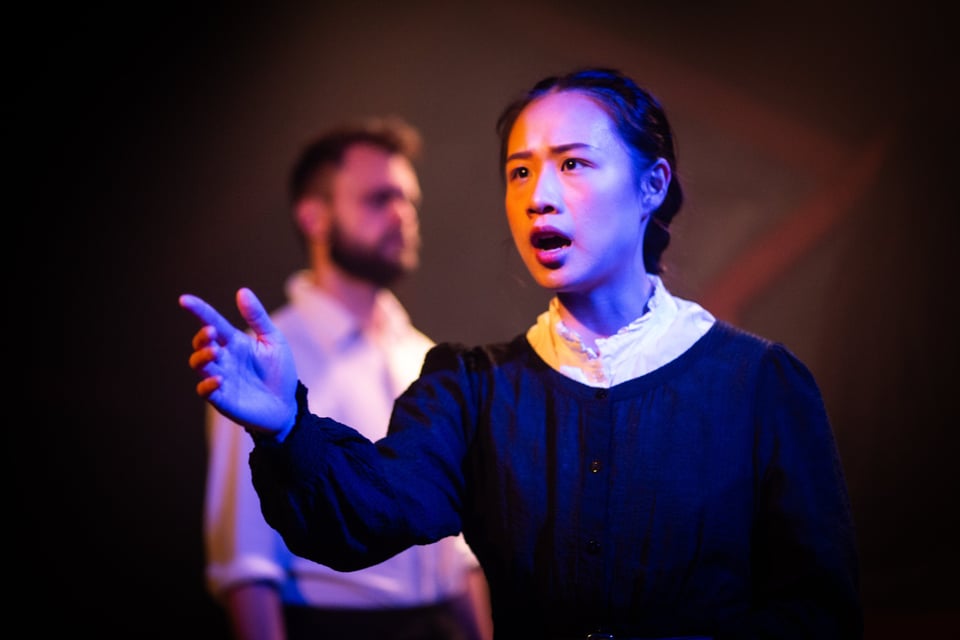 In The Beautiful Future Is Coming: in the foreground, Sabrina Wu (wearing a black Victorian-era dress with a white neck) talks to the audience. George Fletcher, bearded with a white shirt, stands blurred in the background.