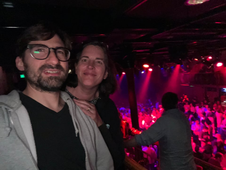 Two people smile at the camera with the concert audience below bathed in red and purple light.