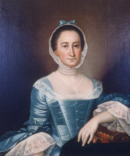 A portrait of a woman with dark hair, wearing a lot of things that are about to be described in the text.