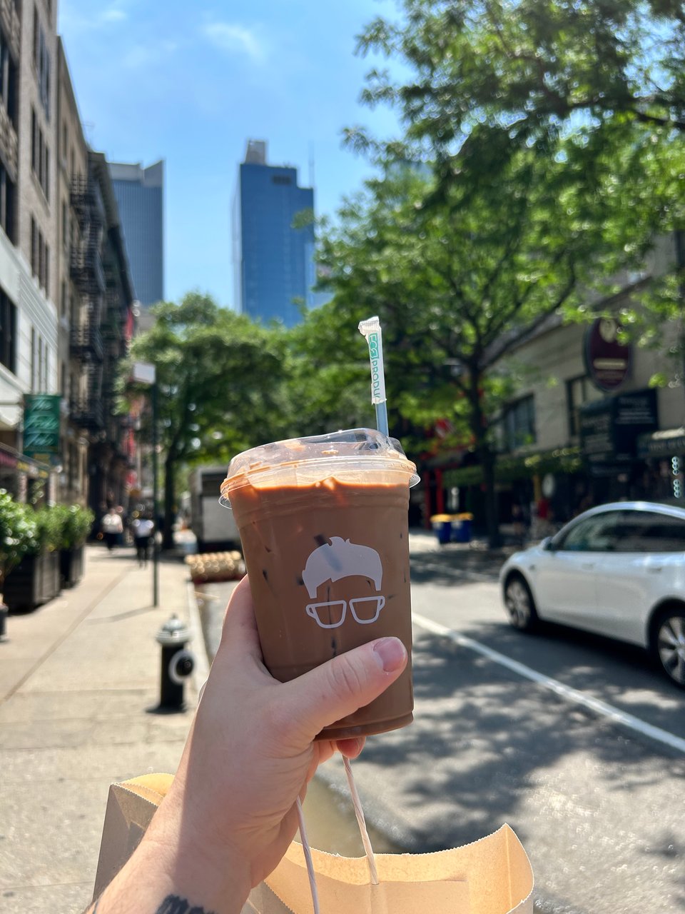 von's hand holding an iced latte in a plastic cup with a blurred background of the new york city skyline