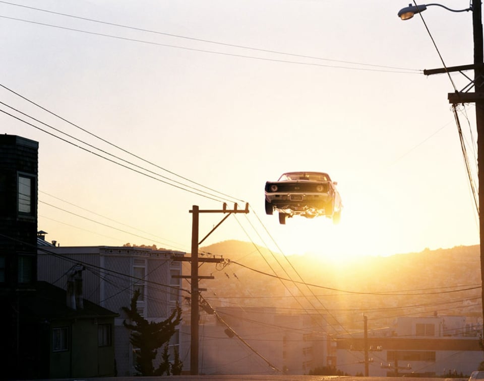 A photograph by Matthew Porter of a car seemingly suspended far above the ground at about the same height as the telephone poles in the picture; the glare of a sunrise behind the car creates golden haze and evokes a peaceful feeling that contrasts with the flying car.